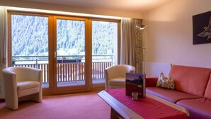 Ein Zimmer mit zwei Sesseln, einem Sofa, einem Tisch mit einem Laptop und einer Tasse sowie einem großen Fenster mit Blick auf eine bewaldete Landschaft.