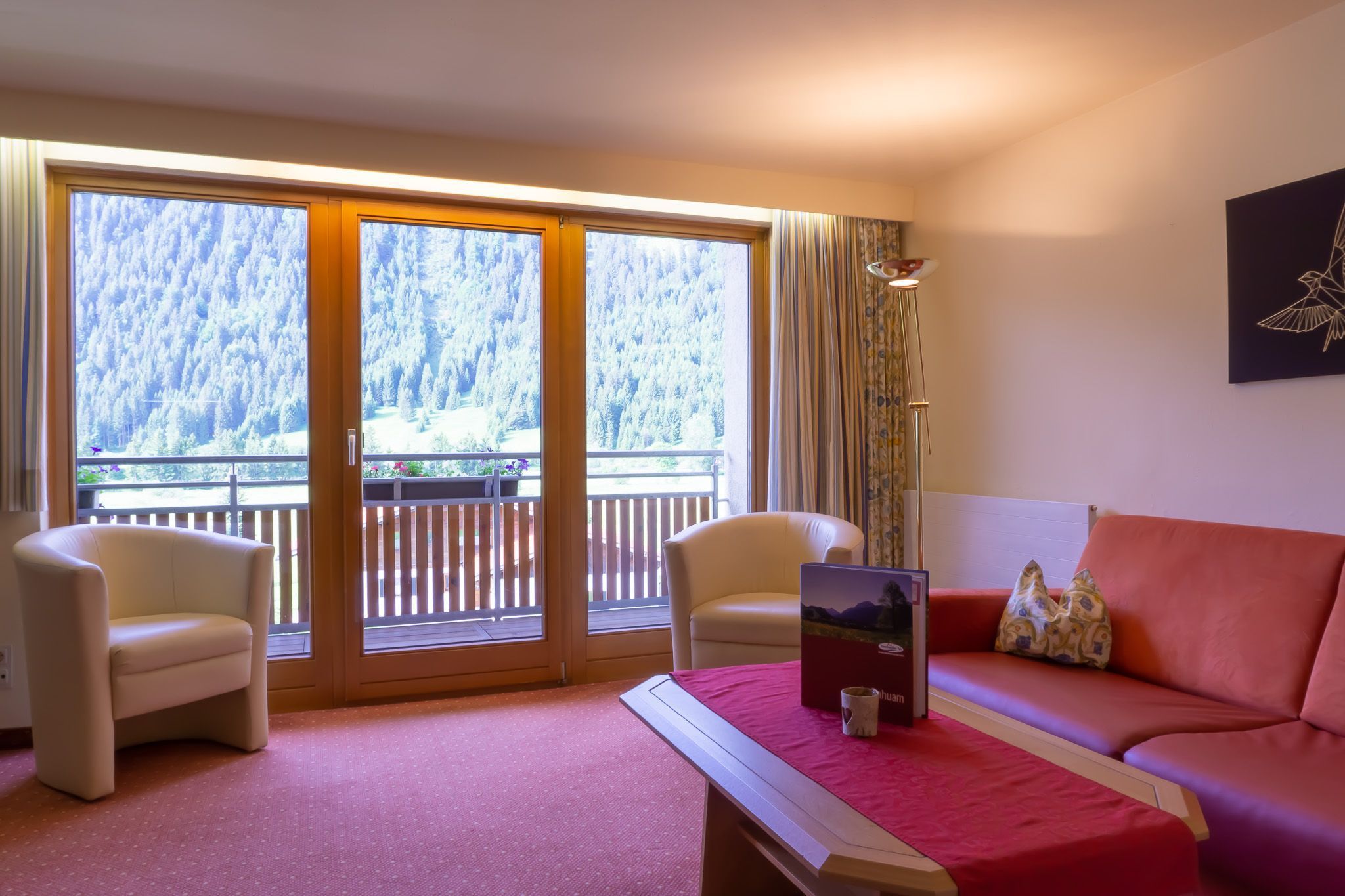 Ein Zimmer mit zwei Sesseln, einem Sofa, einem Tisch mit einem Laptop und einer Tasse sowie einem großen Fenster mit Blick auf eine bewaldete Landschaft.