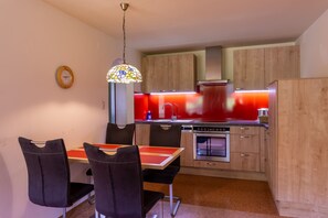Une cuisine compacte avec des meubles en bois, un plan de travail rouge, un four encastré et un coin repas avec des chaises et une table noires.