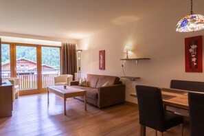 A modern living room with a sofa, coffee table, dining table, chairs, a hanging light fixture, and a view of a building through large windows. - No. 13 Fire lily - HAUS fern (Grän)