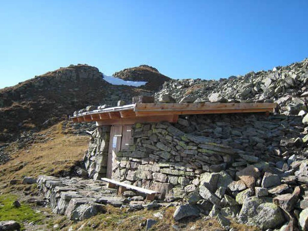 Eine Steinstruktur mit Holzdach, möglicherweise eine Schutzhütte oder Aussichtsplattform, umgeben von Steinen und vereinzelter Vegetation.