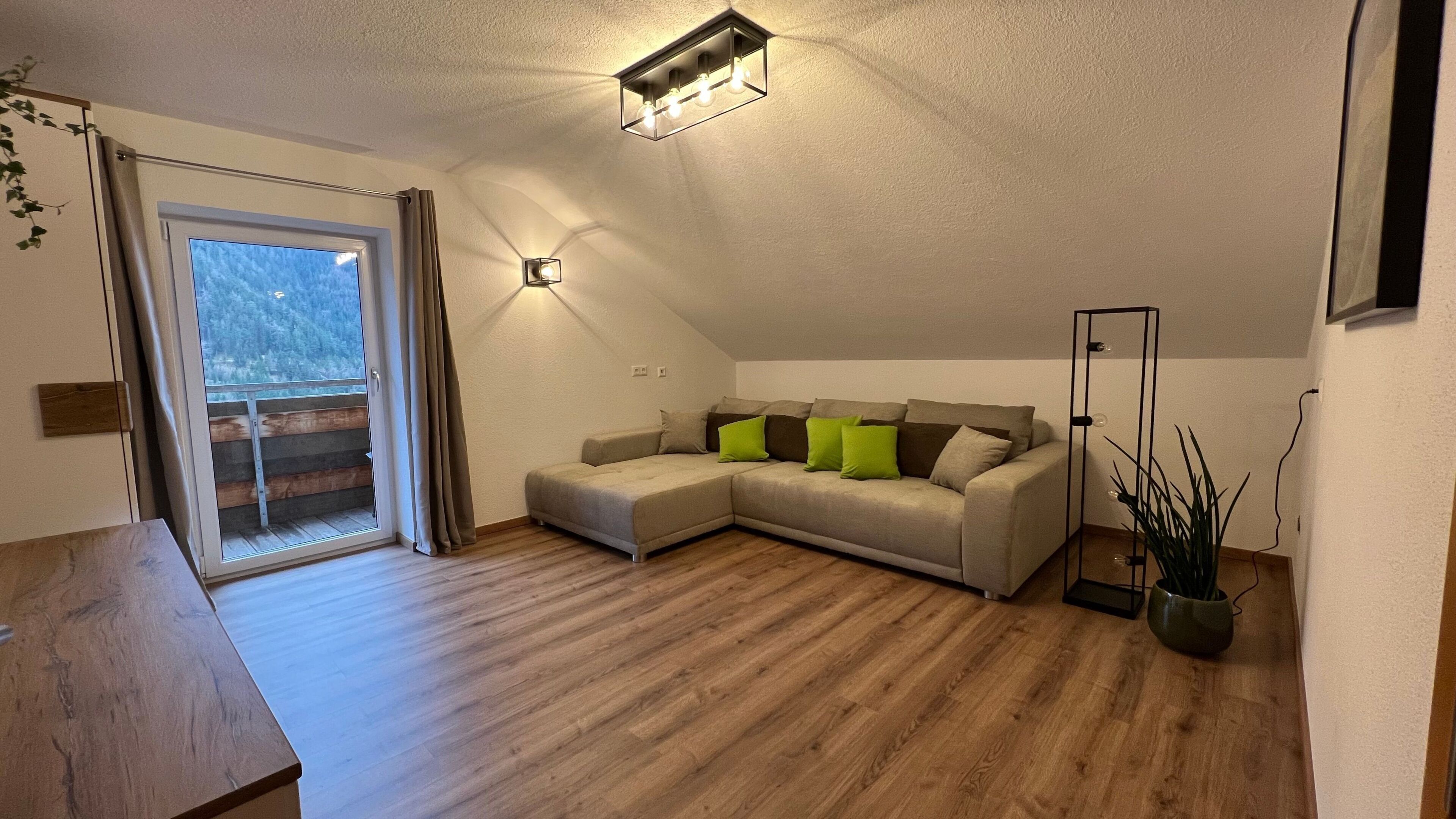 Ein modernes Wohnzimmer mit Holzboden, einem Ecksofa mit Kissen, einer Stehlampe und einer Glastür, die auf einen Balkon führt.