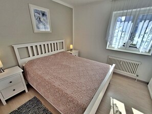 Une chambre à coucher avec un lit, une table de chevet, une lampe, une fenêtre avec des rideaux et un radiateur.