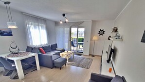Een moderne woonkamer met een bank, een eettafel en een salontafel. Er is een groot raam met gordijnen, een plafondlamp en een wandklok.