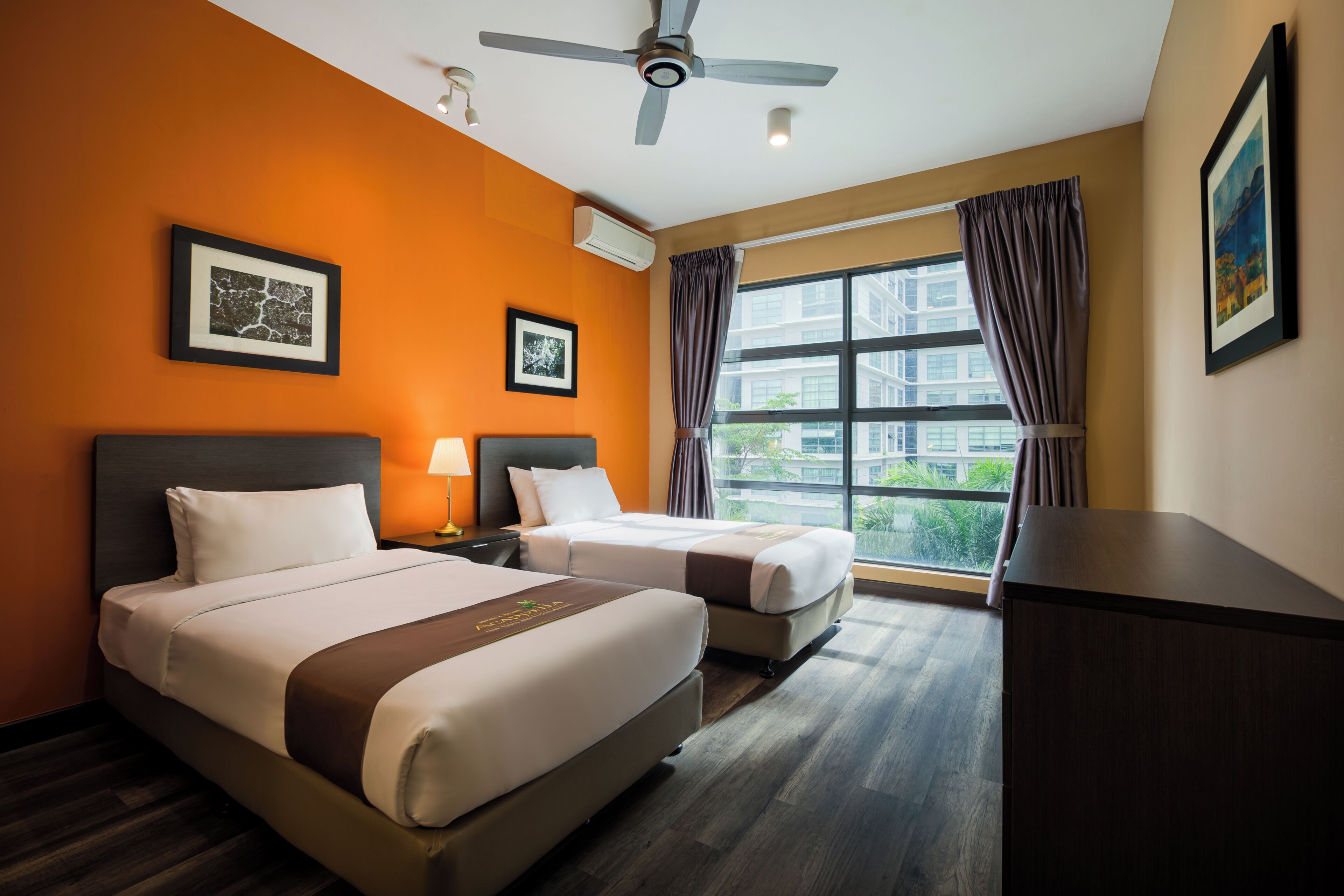Una habitación de hotel con dos camas, ventilador de techo, ventanas amplias con cortinas y cuadros en las paredes.