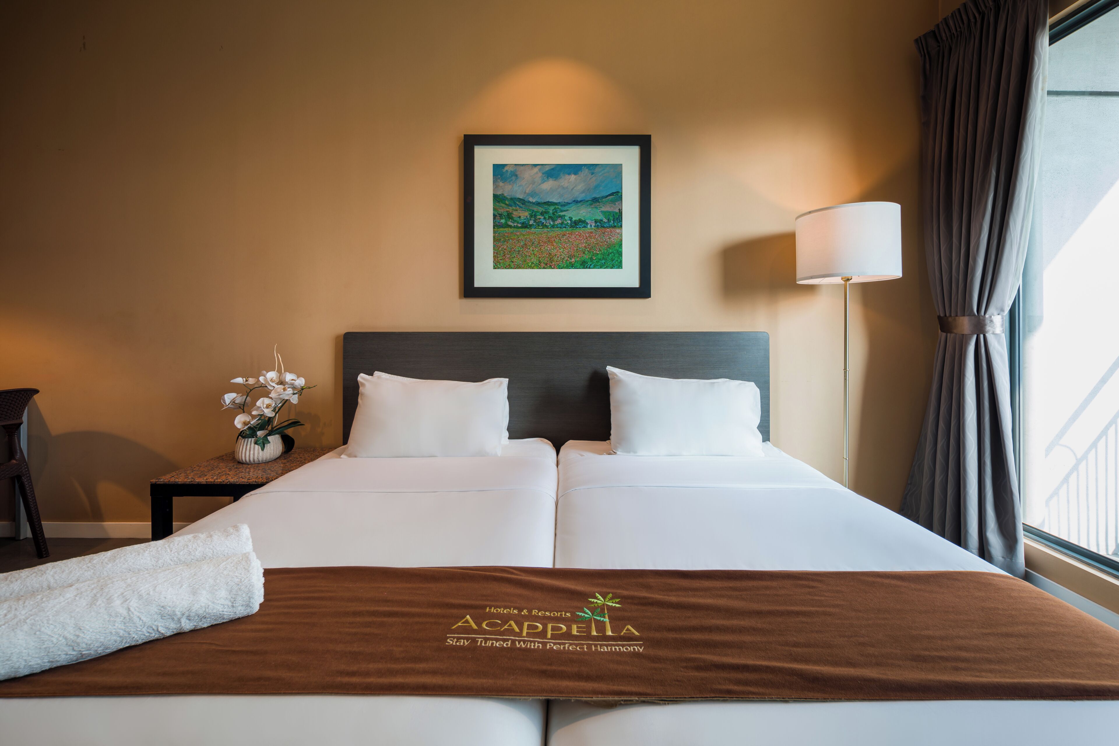 A hotel room with a bed, a framed painting, a lamp, and a window with curtains.