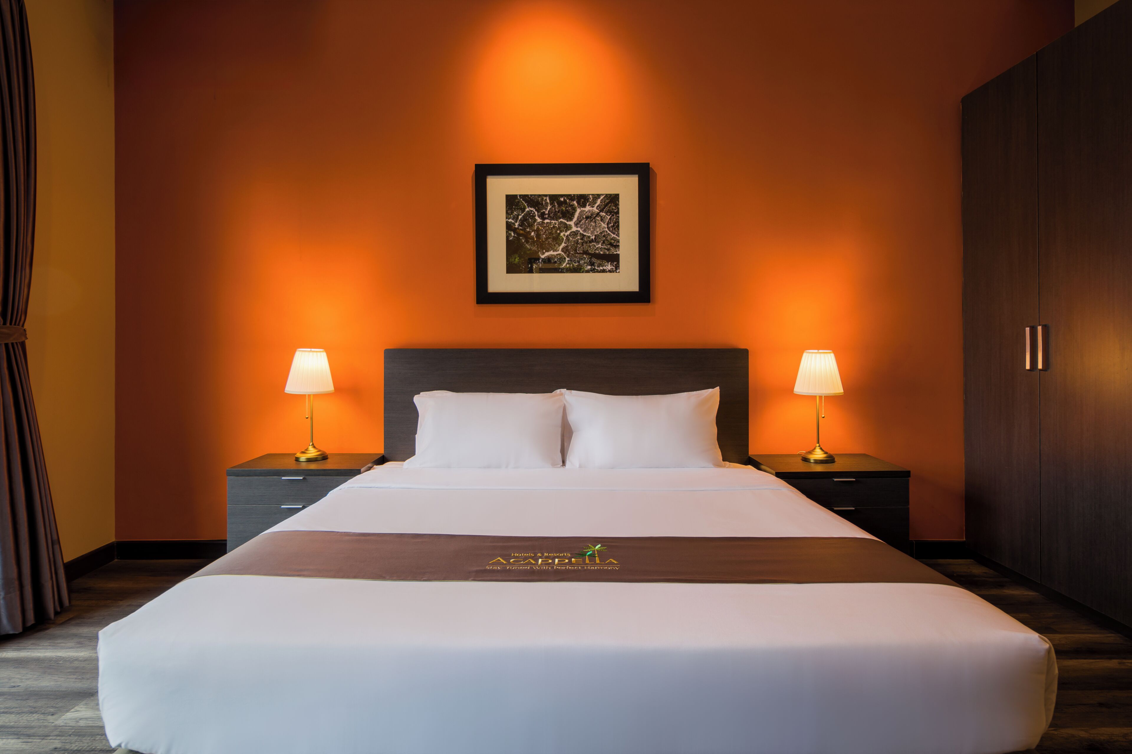 A hotel room with a large bed, two bedside tables with lamps, a framed picture on the wall, and a dark wooden wardrobe.