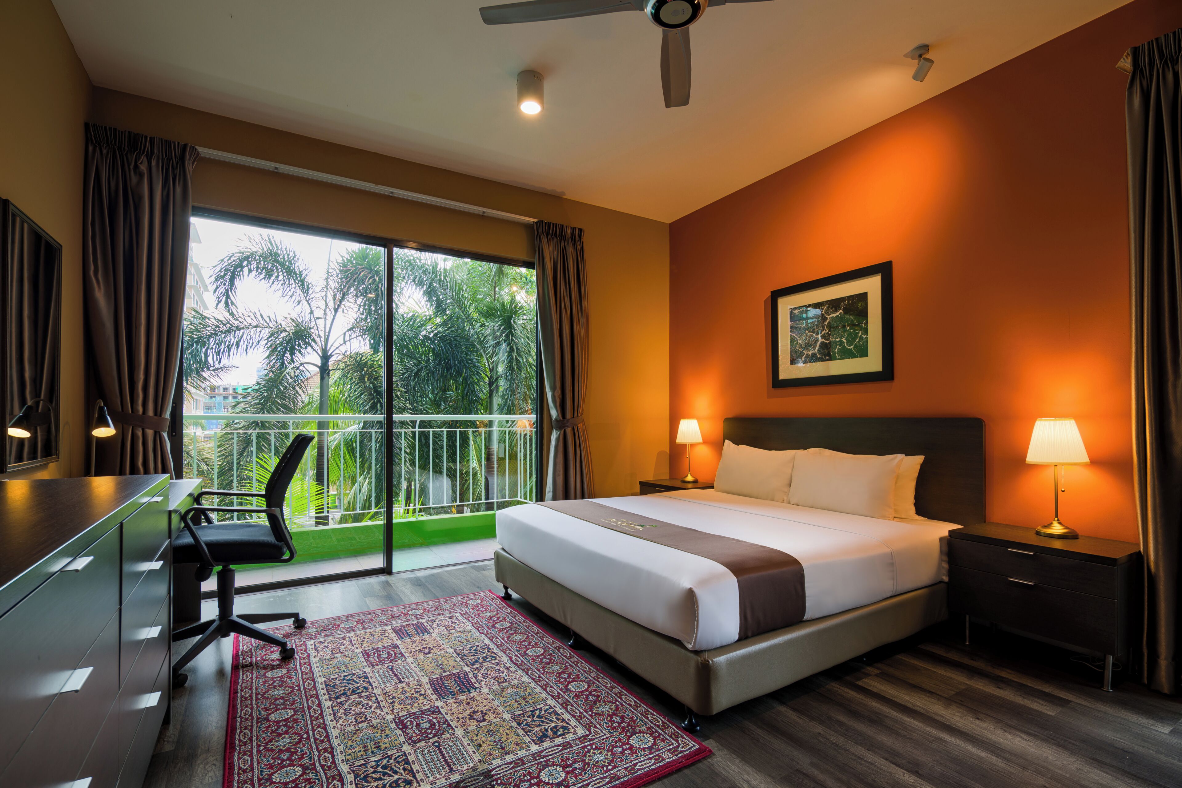 A hotel room with a bed, desk, chair, and a view of palm trees through large windows.