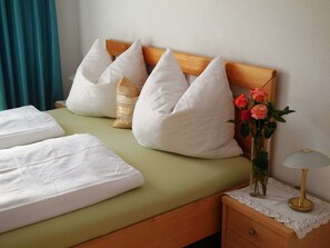 Una cama bien tendida con sábanas blancas, una colcha verde y almohadas decorativas. Hay un florero con flores en la mesita de noche junto a la cama.
