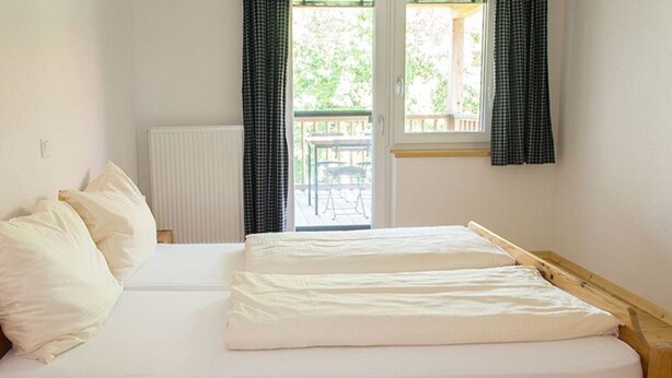 A single bed with a wooden frame, white bedding, and a view of an outdoor deck through a window.