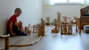 A room with a child playing with wooden blocks, a wicker chair, and a window with curtains.