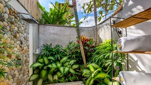 A small, enclosed garden area with a stone wall, lush green plants, a concrete bench, and a towel rack. - Villa Vitari (Seminyak)