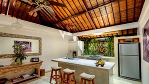 A kitchen with a central island, wooden stools, a ceiling fan, and a stone wall with greenery. - Villa Vitari (Seminyak)