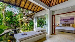 A bedroom with a bed, a small sitting area with a view of a garden and pond, and a painting of a boat on the wall. - Villa Vitari (Seminyak)