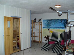 Una habitación con puerta de madera, estante con toallas dobladas, pintura con motivos playeros, silla a rayas y cojín rojo, y un ventilador de techo.