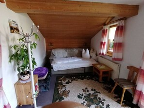 A cozy bedroom with a bed, a wooden ceiling, a window with curtains, a small table, a chair, and a plant.