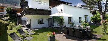 A modern outdoor dining area with a glass table and chairs, surrounded by a well-maintained garden and a white building with a sign reading "GASTEHAUS ZINNER".
