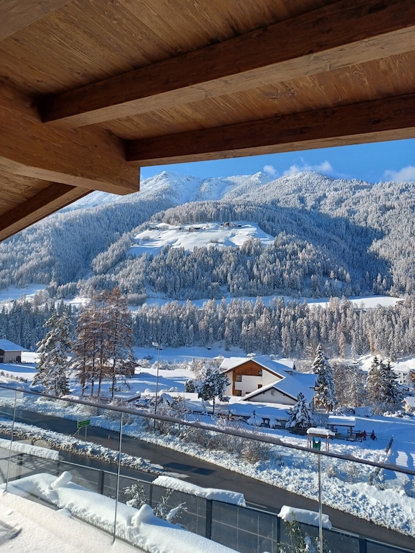A view from a wooden balcony overlooking a snow-covered landscape with mountains in the background and a road in the foreground.