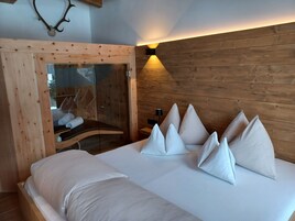 Un dormitorio moderno con cabecera de madera, baño con mampara de vidrio y cama con ropa y almohadas blancas.
