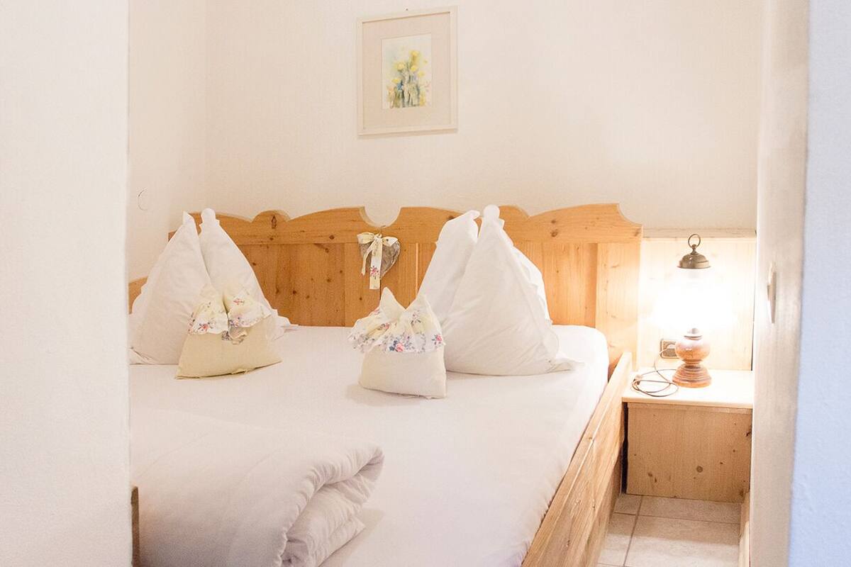 Een netjes opgemaakt bed met bloemenkussens, een houten hoofdbord, een nachtkastje met een lamp en een lijst met een afbeelding aan de muur.