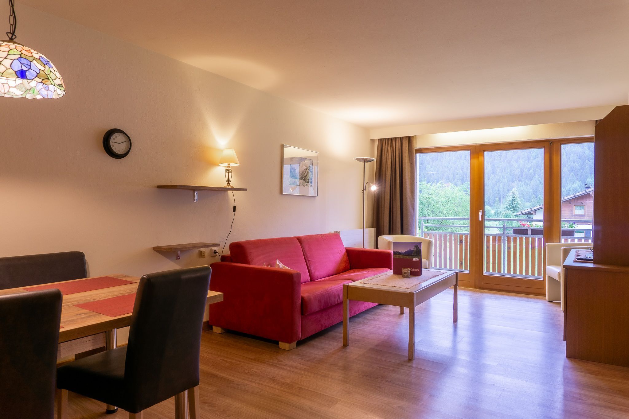 Ein Wohnzimmer mit rotem Sofa, Esstisch aus Holz und Stühlen, einem Couchtisch, einer Uhr, einer Lampe und einem großen Fenster mit Blick auf Bäume und Gebäude.