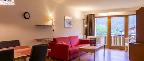 Una sala de estar con un sofá rojo, una mesa de comedor de madera y sillas, una mesa auxiliar, un reloj, una lámpara y un ventanal con vistas a árboles y edificios.