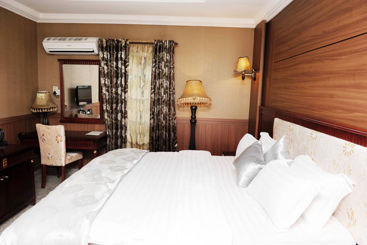 A hotel room with a large bed, a desk, a chair, a lamp, and an air conditioning unit.