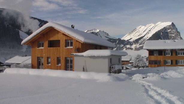 A wooden chalet with a snow-covered roof, surrounded by a snowy landscape and mountains. - Apartment Simone - Apartment Simone (Schoppernau)