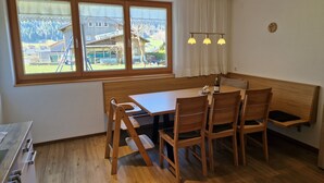 A dining area with a wooden table and chairs, a bench, and a view of a house and mountains through large windows. - Apartment Simone - Apartment Simone (Schoppernau)