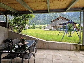 A covered patio with a table and chairs, a swing set, and a view of a green field and houses in the distance. - Apartment Simone - Apartment Simone (Schoppernau)