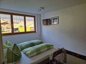 A room with a bed, a window with a view of a house and trees, a wall-mounted picture, and a ceiling light fixture. - Apartment Simone - Apartment Simone (Schoppernau)