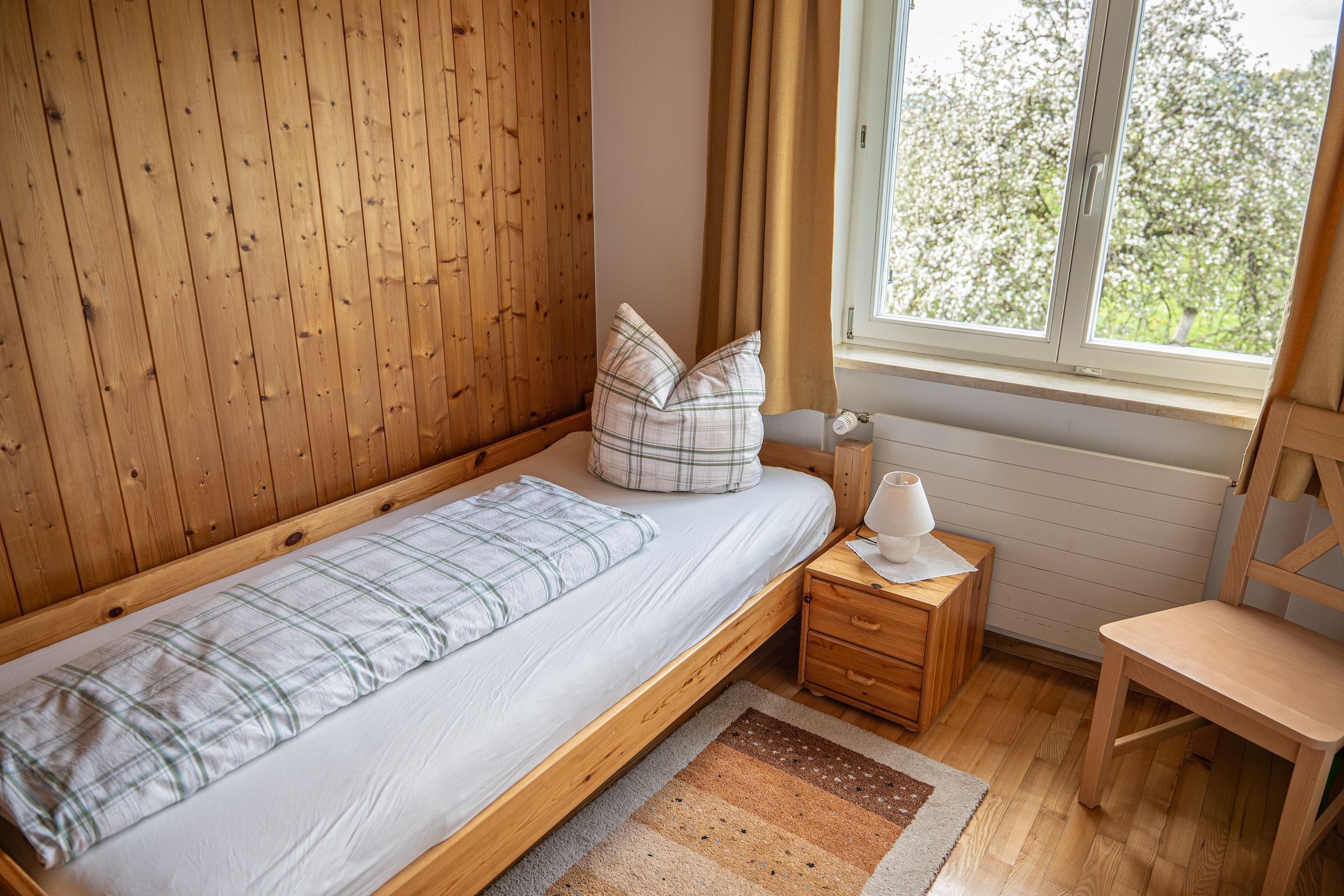 Un lit simple avec une couverture à carreaux et des oreillers, une table de chevet avec une lampe, une chaise et une fenêtre avec des rideaux.