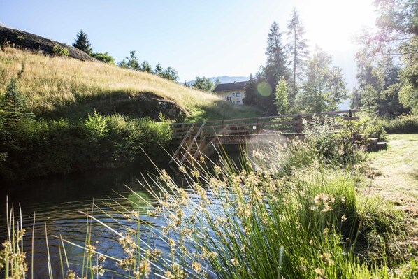 A river with a wooden bridge, surrounded by greenery and a house in the background.