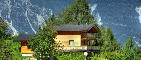 A wooden cabin with a balcony, surrounded by lush greenery and mountains.