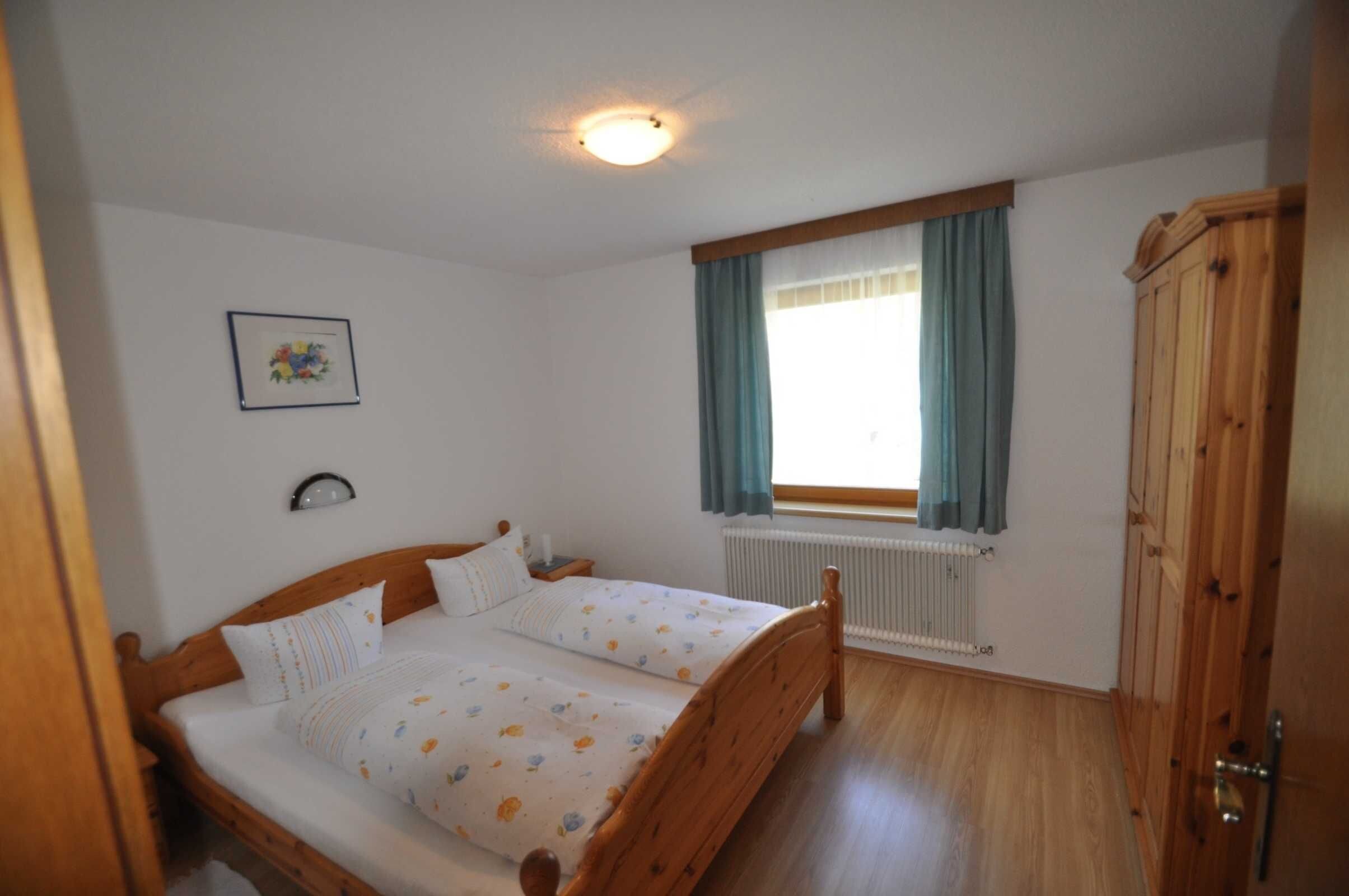 Une chambre avec un lit en bois, un radiateur, une fenêtre avec des rideaux, un tableau encadré au mur et une armoire.