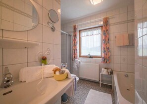 Un baño con bañera, lavabo con espejo, ventana con cortinas y un perchero para toallas.
