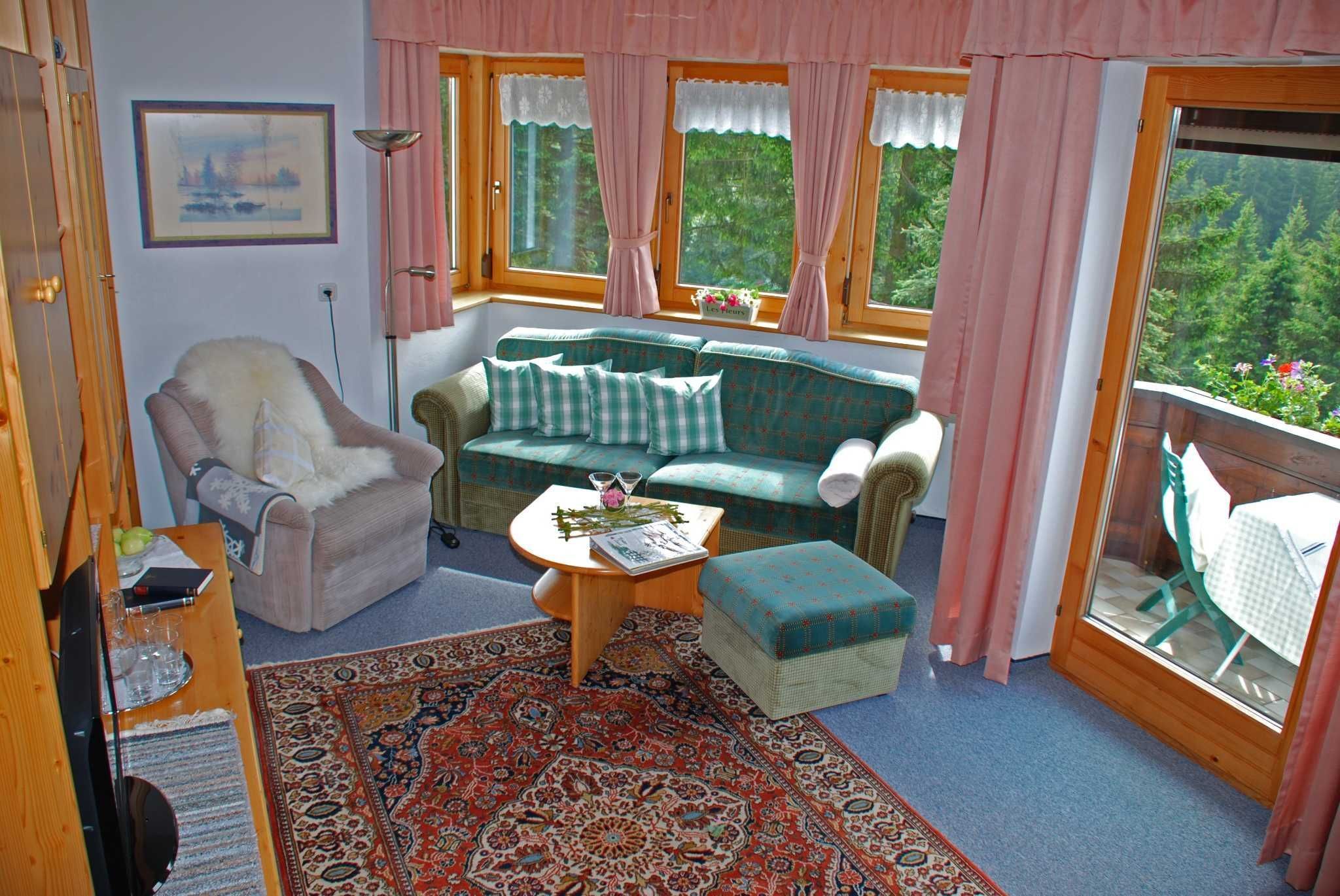 Ein gemütliches Zimmer mit einer Couch, einem Sessel und einem Hocker, einem kleinen Tisch mit einem Buch und Gläsern sowie einem großen Fenster mit Vorhängen.