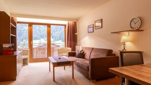 A room with a sofa, a table, a chair, a desk, a lamp, a clock, and a view of a balcony with a forest. - No. 8 Bergaster - HAUS fern (Grän)