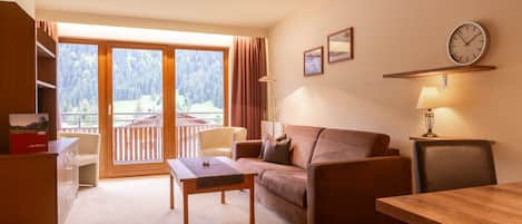Una habitación con sofá, una mesa, una silla, un escritorio, una lámpara, un reloj y vistas a un balcón con bosque.
