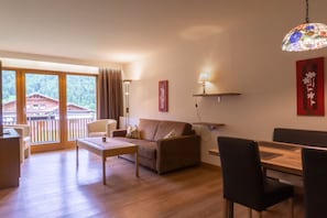 Ein modernes Wohnzimmer mit einer Couch, einem Couchtisch, einem Esstisch, Stühlen, einer Pendelleuchte und Blick auf ein Gebäude durch große Fenster.