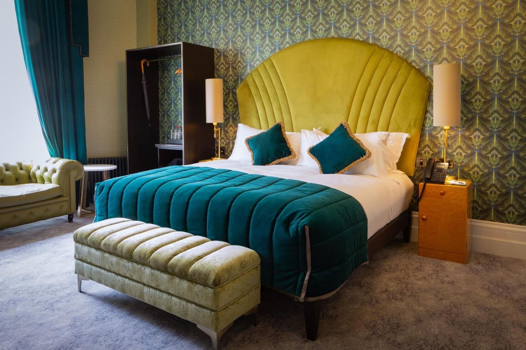 Habitación de hotel con una cama grande, un cabecero tapizado en verde, un banco, una mesita de noche con una lámpara y un papel tapiz estampado.