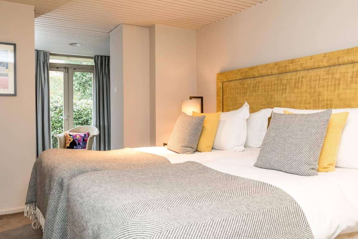 A hotel room with a large bed, a headboard, a nightstand with a lamp, and a view of greenery through a window.