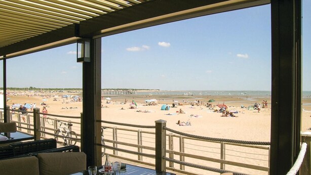 Un restaurante frente a la playa con terraza y vistas a una playa concurrida.