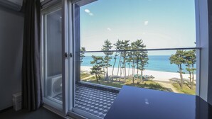 A room with a large window overlooking a beach with trees and the sea.