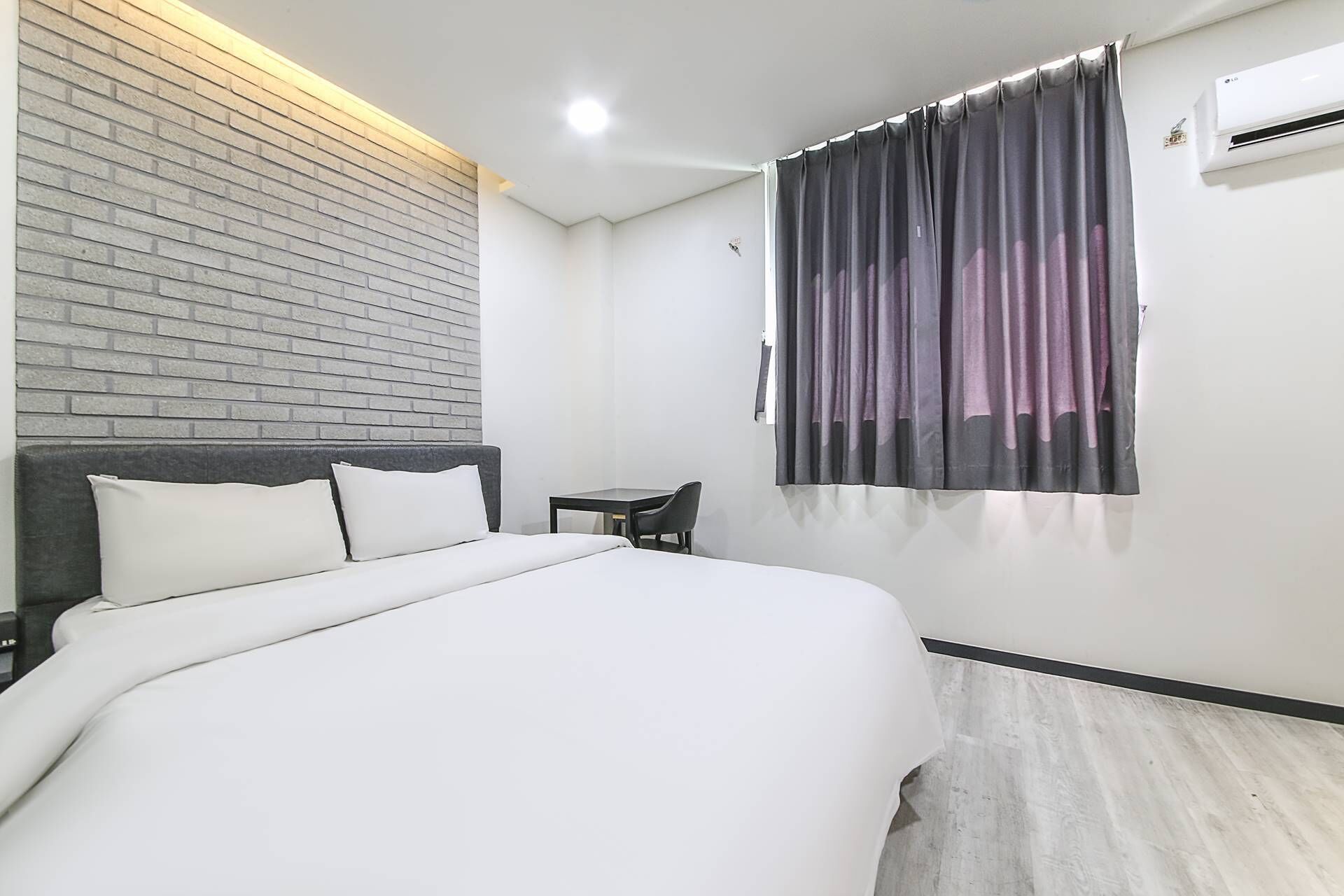 A hotel room with a bed, a small table, a chair, a window with curtains, a brick wall, and an air conditioning unit.
