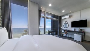 A modern hotel room with a large bed, a desk, a chair, a TV, and a view of the sea.