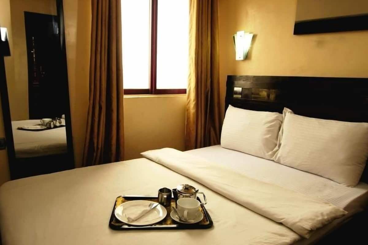 A hotel room with a bed, a tray with breakfast items, a window with curtains, and a wall-mounted light fixture.