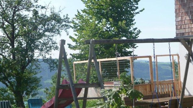 Een houten terras met een schommelset, een glijbaan en een hangstoel.