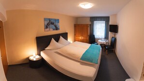 Quarto de hotel com cama, luminária de cabeceira, uma mesinha com cadeira, televisão na parede e uma janela com cortinas.