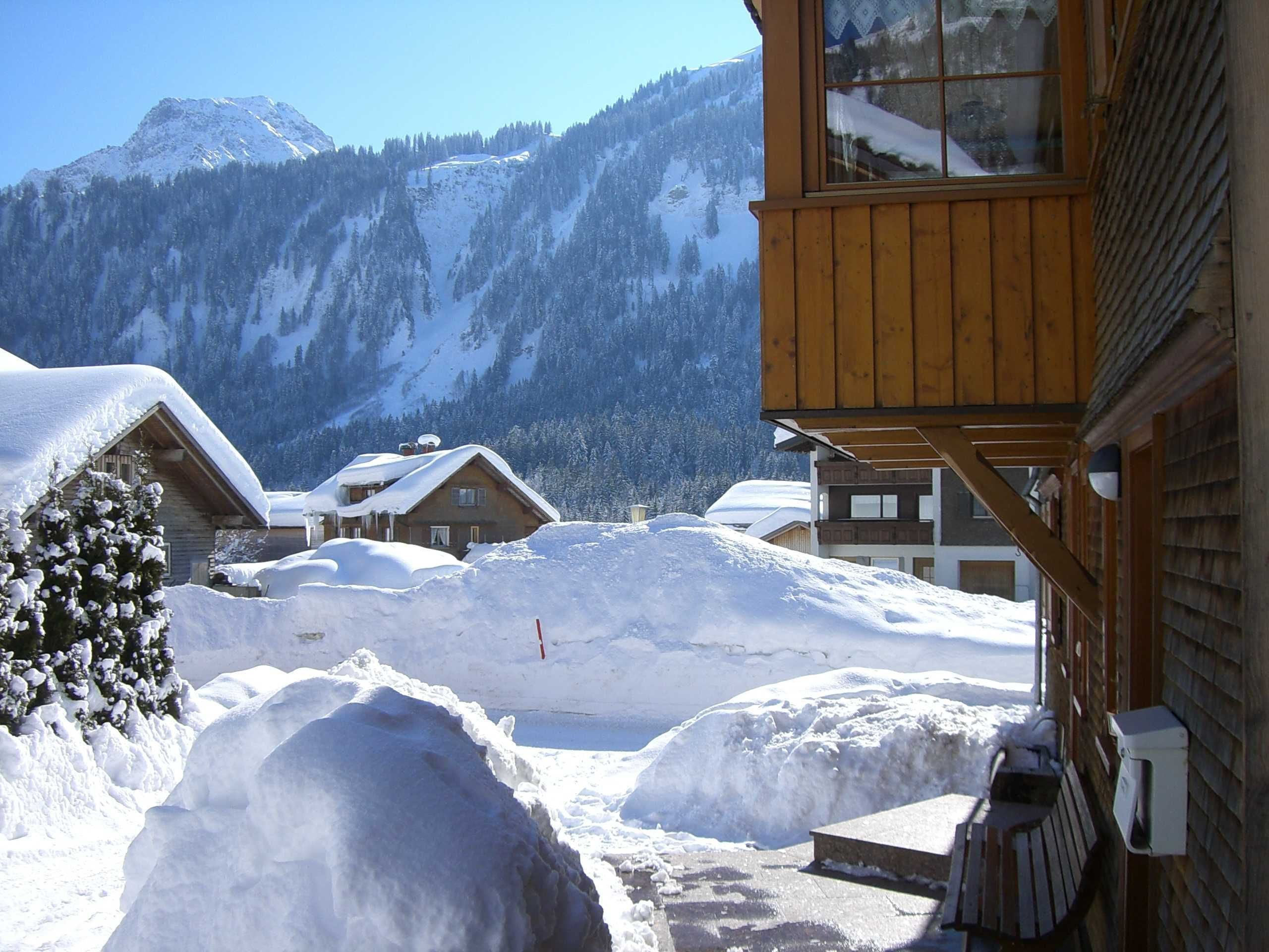 Een houten chalet met een met sneeuw bedekte dak, een houten terras en uitzicht op een besneeerd berglandschap.